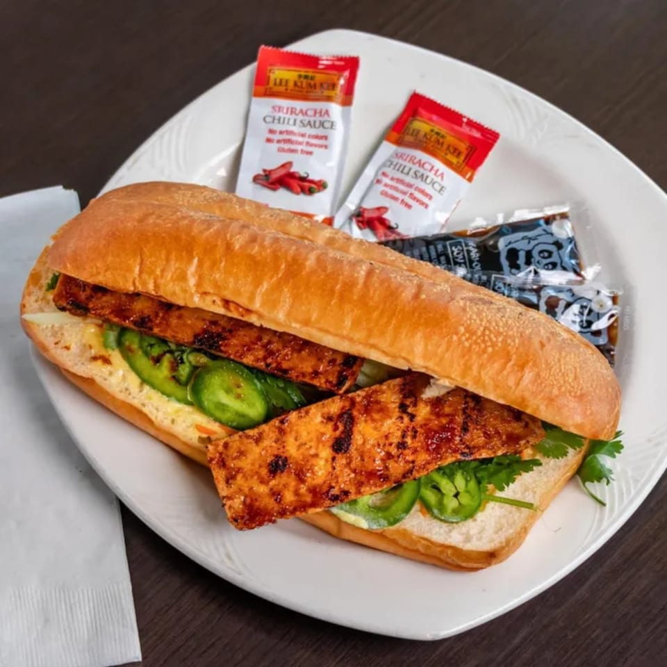 A Vietnamese banh mi sandwich with grilled meat, jalapenos, and cilantro, served with chili sauce packets.