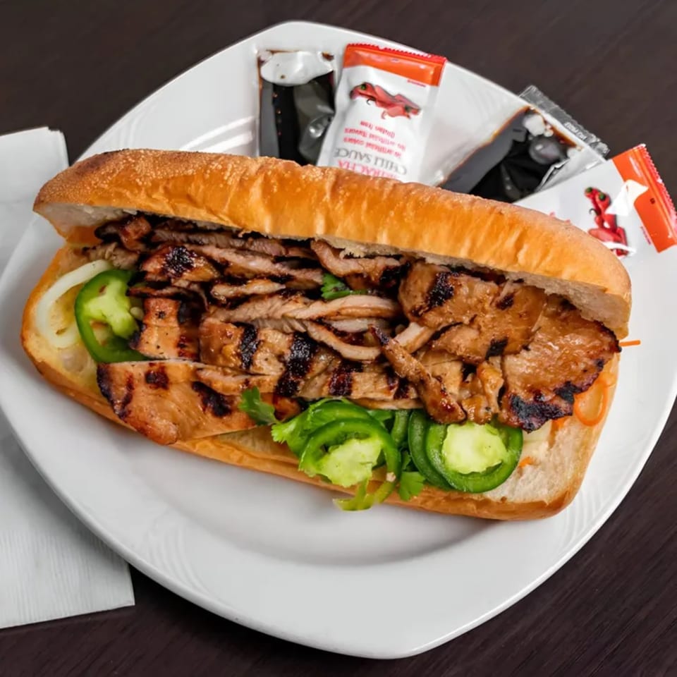 Grilled pork banh mi sandwich with jalapeños, onions, and cilantro on a white plate with sauces.